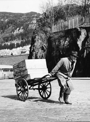 Svart-hvitt bildet av mann med fiskekasser på kjærre.